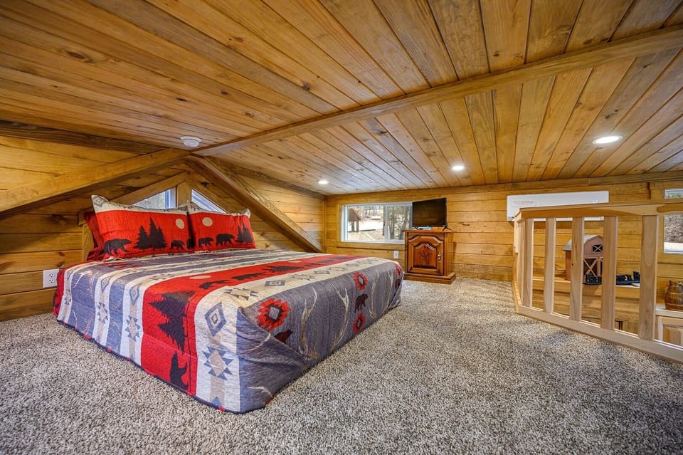 A cozy loft bedroom with a wooden ceiling and walls, carpeted floor, a bed with a bear-themed quilt, a small window, a TV, and a wooden railing by the stairs.