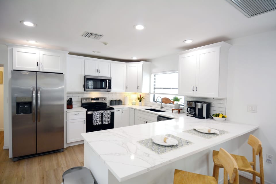 Modern kitchen with white cabinets, stainless steel appliances including a refrigerator and oven, marble countertops, a large window, and wooden flooring. A plant is placed on the counter.