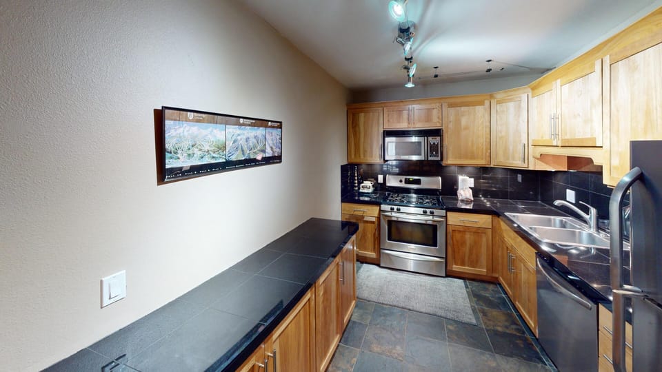A modern kitchen with wooden cabinets, stainless steel appliances, black countertops, a double sink, and a wall-mounted picture.