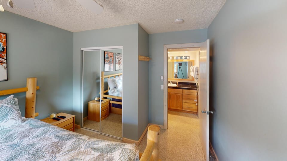 A bedroom with a bed, mirrored closet doors, and a nightstand, leading to an adjacent bathroom with a wooden vanity and a large mirror.