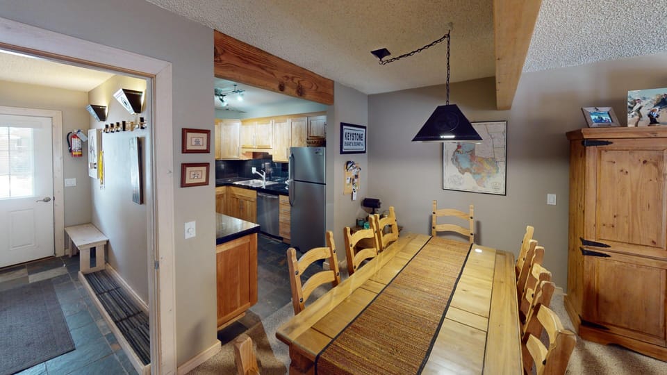 A dining area with a wooden table and chairs, adjacent to a kitchen with light wood cabinets and stainless steel appliances. A map, photos, and a cupboard decorate the space.