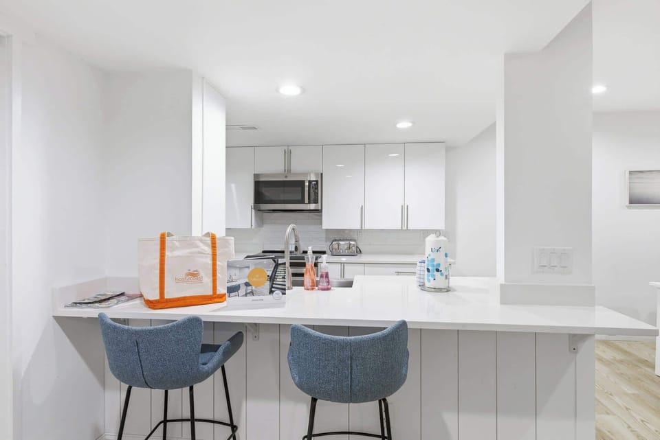Crisp white cabinetry and quartz counters create a bright, airy culinary space perfect for slow mornings. Azure barstools add a pop of coastal color to this polished kitchen, where every detail feels fresh and impeccably clean.