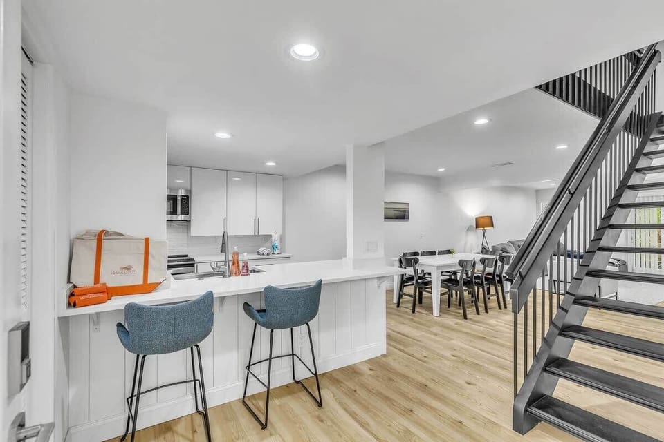 A striking black staircase anchors this airy, open layout, where crisp white walls meet soft wood floors. Bright recessed lighting and a communal dining space create a clean, modern atmosphere perfect for gathering after a day of adventure.