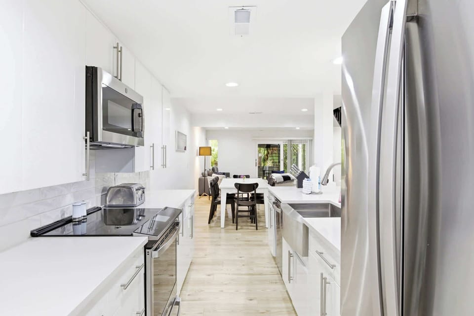 Stainless steel appliances and snowy surfaces create a culinary space that feels airy and organized. Soft overhead glow illuminates the open galley, leading your eyes toward the shared dining area and the leafy views beyond.