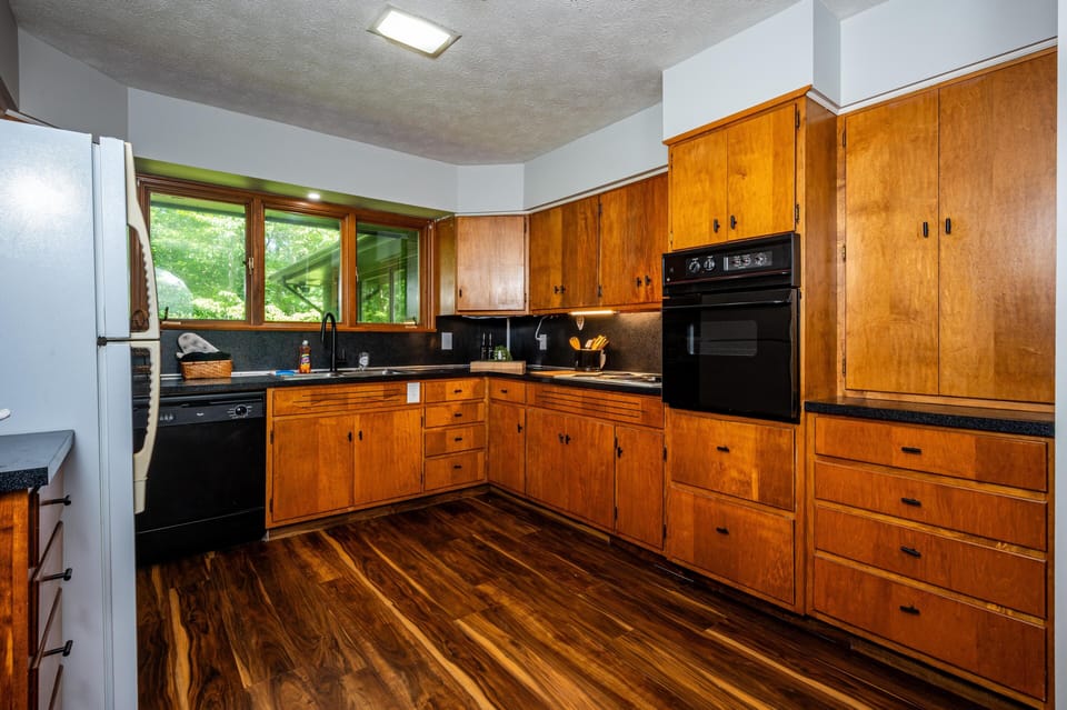 The kitchen features honey-toned wood cabinets, black appliances, and a wide window overlooking lush greenery.