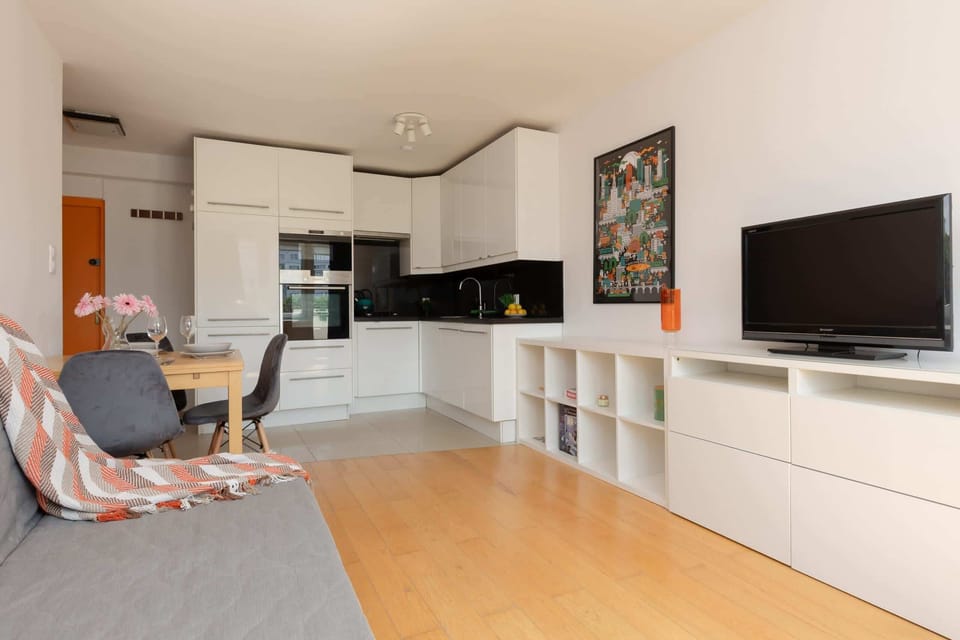 A modern living room with a TV, minimalist furniture, and an open-plan layout connecting to the kitchen.

