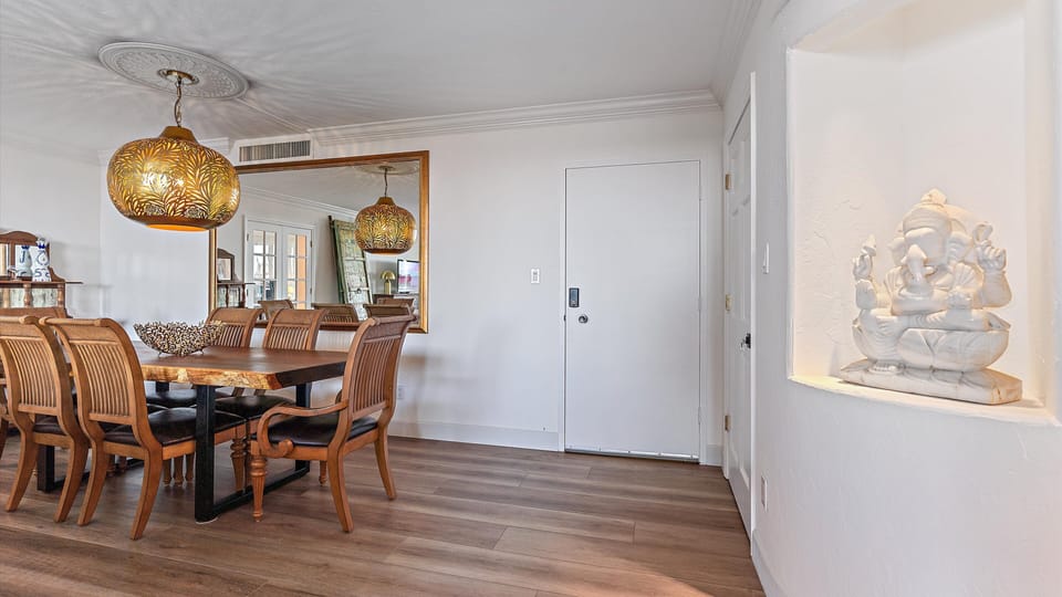 A dining room with a wooden table and chairs, pendant lights with decorative shades, a large wall mirror, and a niche displaying a white statue. The walls and floor are different shades of wood.