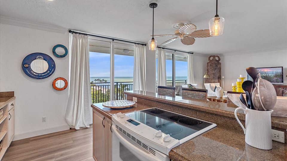 Modern kitchen with a view of the ocean, featuring a stove, countertop, utensils in a holder, and various wall decorations. Sliding doors open to a balcony with a scenic seascape.