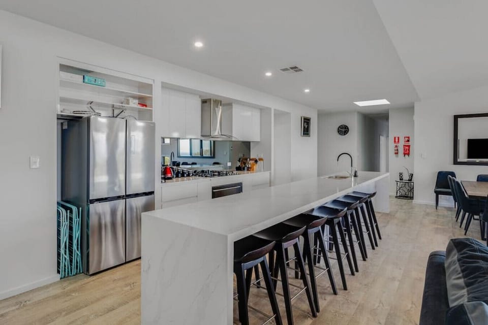 A modern, open-plan kitchen and living area. It features a sleek stainless steel refrigerator, a long marble kitchen island with black stools, and a dining table with chairs in the background. The space has a contemporary design with white cabinetry and a