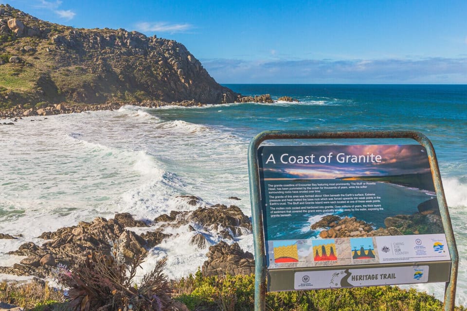 A scenic coastal view with waves crashing against rocky shores by the ocean. There is a sign titled `A Coast of Granite` providing information about the granite coast, indicating it is part of a heritage trail.