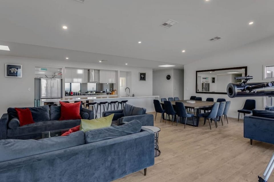 A modern, spacious living area with a combination of a cozy seating space featuring dark gray sofas with red and green cushions, and a dining area with a wooden table and blue chairs. The room has a clean, minimalist design with white walls and ceiling, a