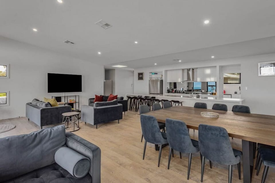 A modern, open-plan living and dining area with a sleek design. It features a large flat-screen TV on a white wall, comfortable gray sofas with colorful cushions, and a long wooden dining table surrounded by gray chairs. The space also includes a well-equ