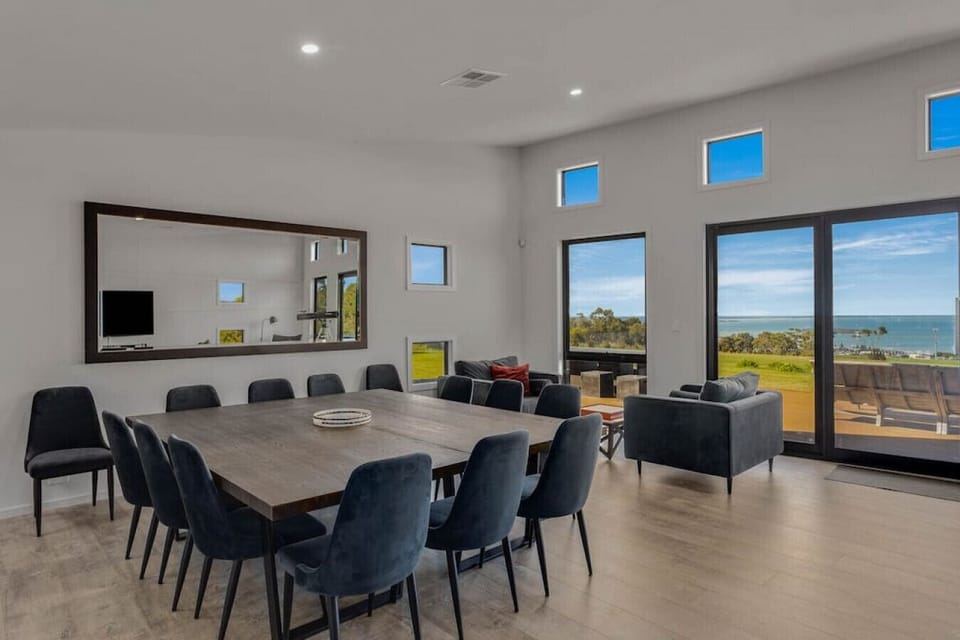 A modern, spacious dining and living area with a large wooden dining table surrounded by dark chairs. The room features large windows offering a scenic view of the ocean and greenery outside, along with a comfortable seating area and a large mirror on the
