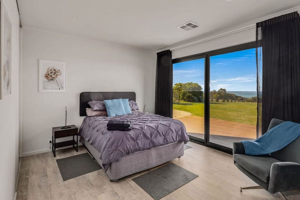 A modern, cozy bedroom with a large sliding glass door that offers a scenic view of a grassy area and a body of water under a clear sky. The room features a bed with a purple quilt and blue pillows, a bedside table, a framed picture on the wall, and a com
