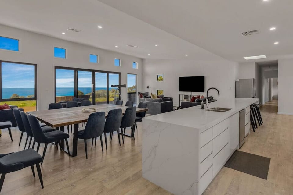 A modern, open-plan living and dining area with a large dining table and dark chairs. The space features a sleek white kitchen counter and a sink, along with a lounge area that includes a television. Large windows offer a stunning view of the ocean, enhan