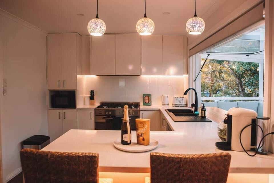Kitchen | With a pristine white stone benchtop illuminated by elegant LED & Pendant lighting; and a large window opening up to the deck, this kitchen offers a harmonious connection between indoor and outdoor living.