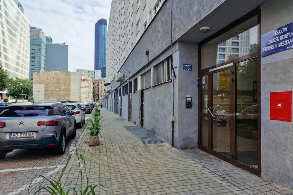 A parking area along the side of a gray building, surrounded by potted plants and urban greenery, leading to the building entrance.
