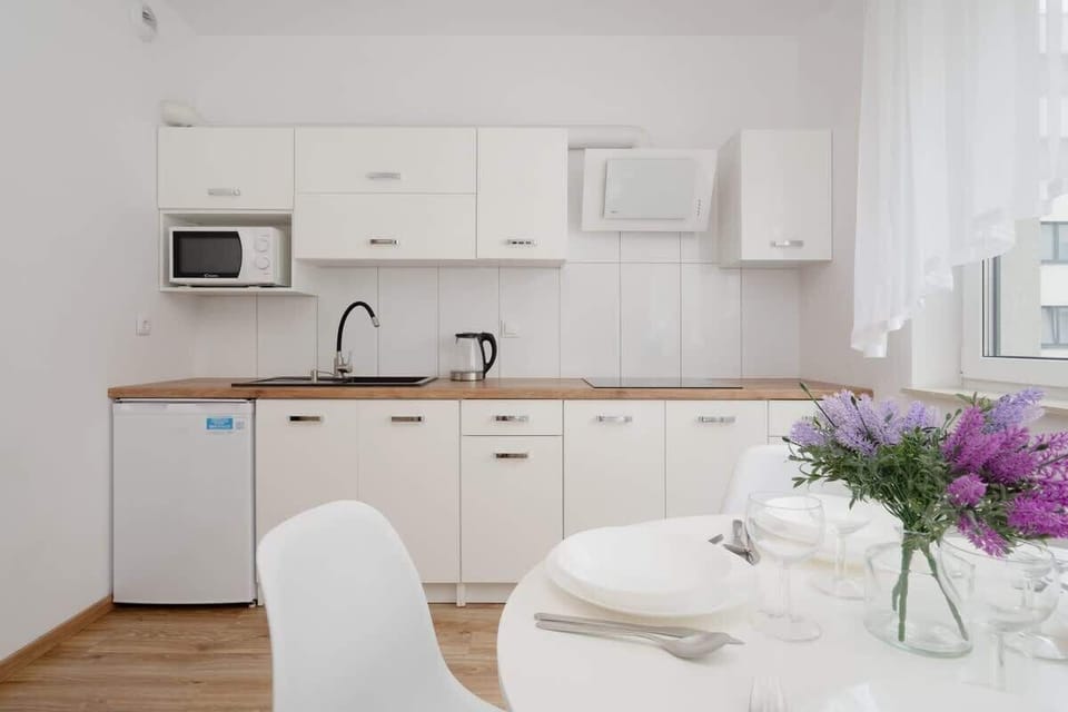 A compact kitchen with white cabinets, a microwave, and a round dining table for two, complemented by a vase of fresh flowers.


