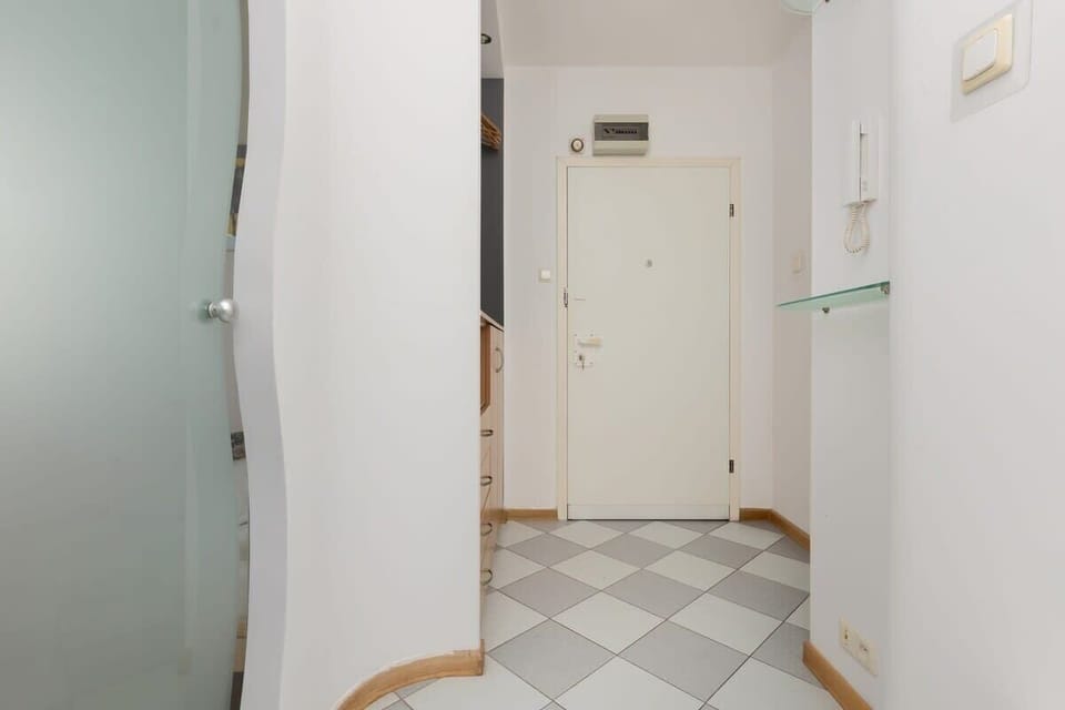 A hallway with light walls, tiled flooring, and a white door leading into the main apartment area, designed to feel clean and welcoming.

