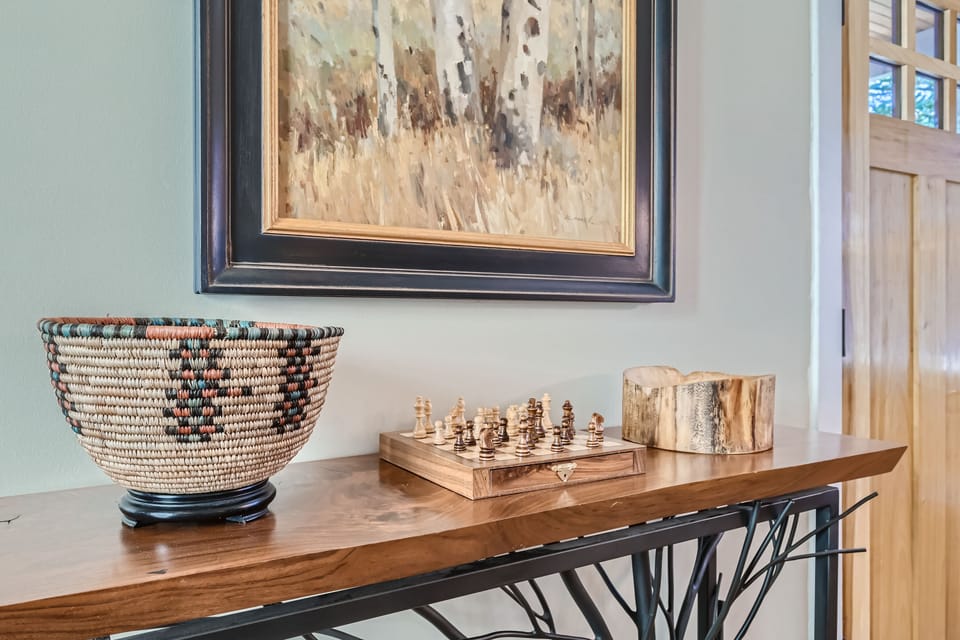 A wooden console table holds a woven basket, a small wooden chess set, and a wooden container. A framed painting of trees hangs on the wall above. The door is partially visible on the right.