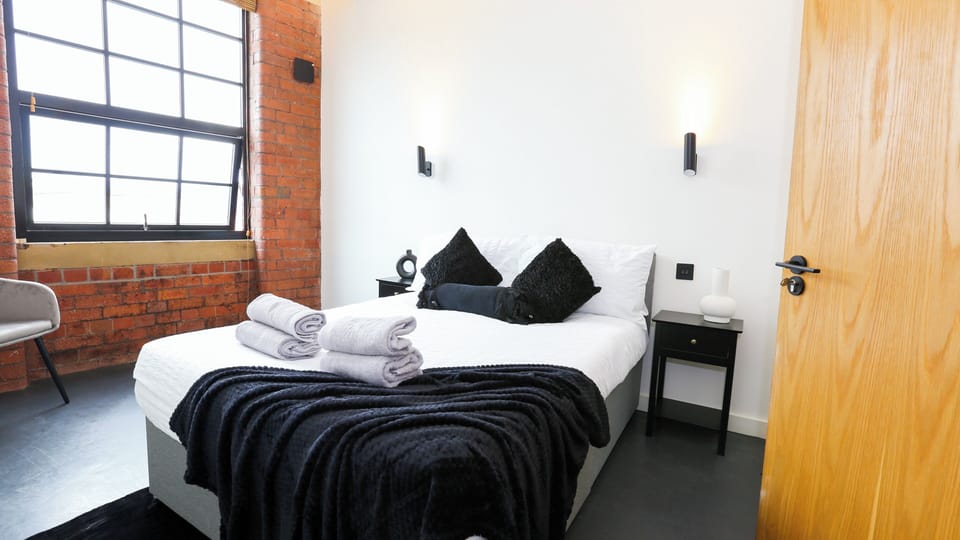 Double bedroom with black cushions, white bedding, exposed red brick feature wall, and tall window – The Nichols Bldg, Sheffield