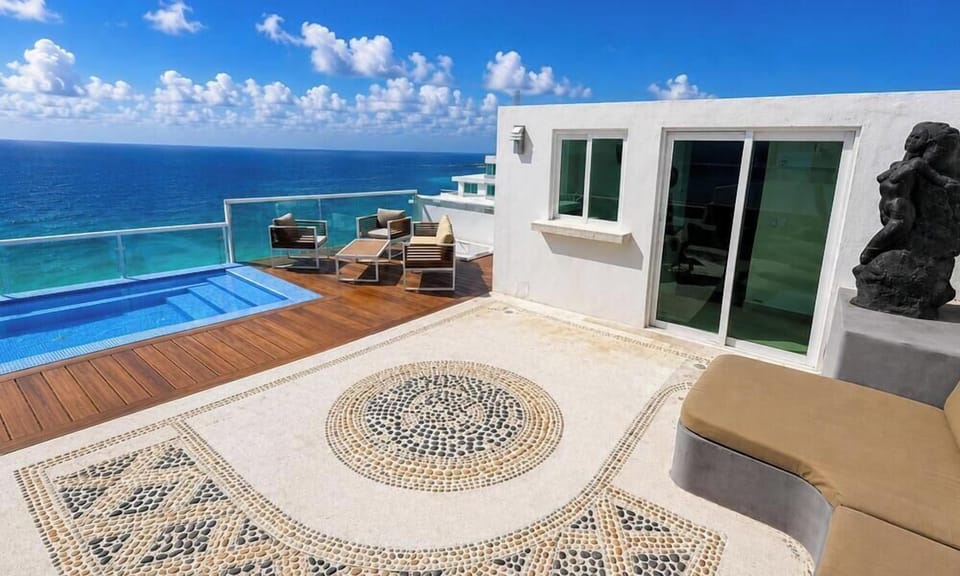 Sunlit terrace with circular pebble mosaic, tan cushioned concrete bench, sliding glass doors, and ocean views.