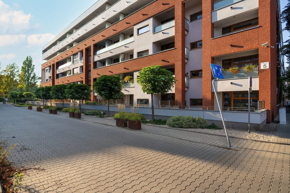 The exterior of an apartment complex, showcasing a well-maintained entrance and surrounding greenery with parking available.