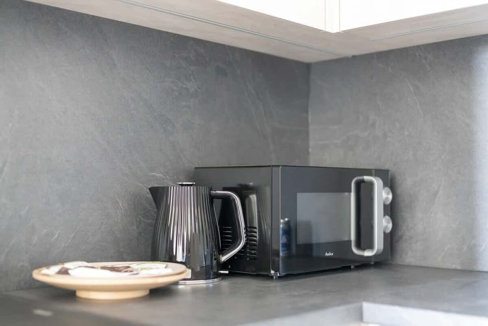 A modern kitchen corner featuring a microwave, a kettle, and minimalist decor, with a sleek gray backsplash.
