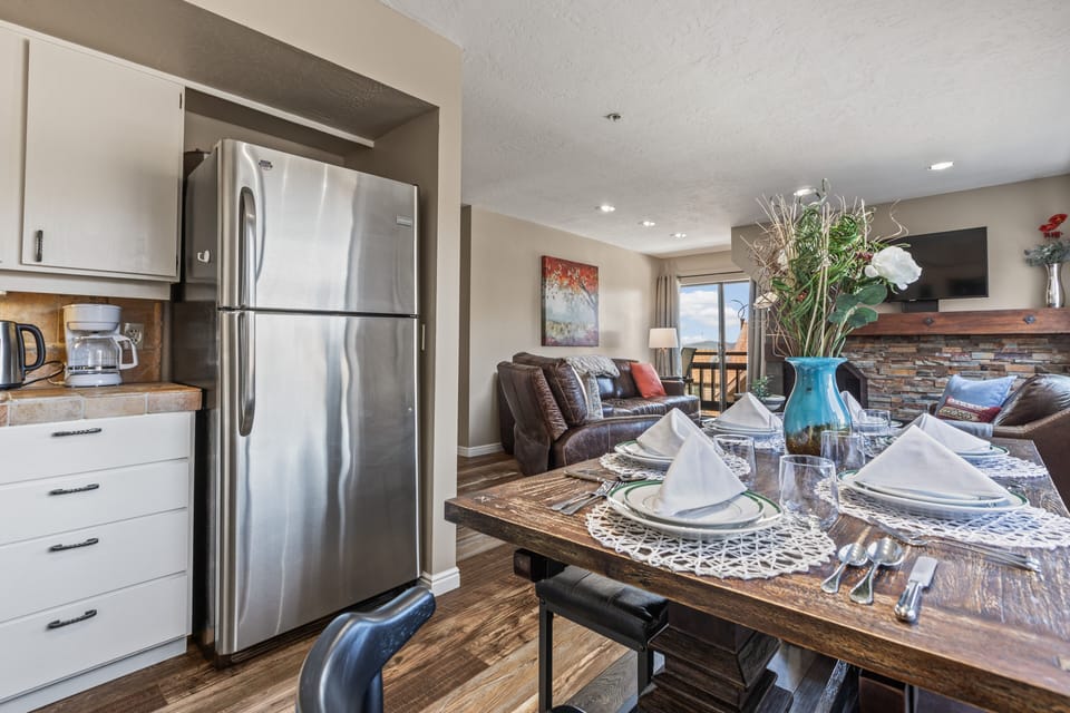 A modern kitchen and dining area with a stainless steel refrigerator, white cabinets, a dining table set for four, and a living room with a brown leather sofa and a fireplace in the background.