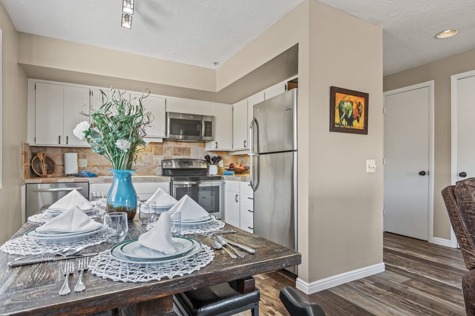 A kitchen with white cabinets and stainless steel appliances. A dining table is set for four with white napkins, plates, and lace doilies. A blue vase with flowers is the centerpiece on the table.