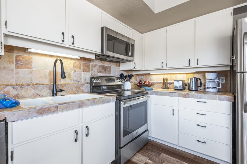 A modern kitchen with white cabinets, stainless steel oven, microwave, and refrigerator, a tile backsplash, black faucet, and small appliances on the counter.