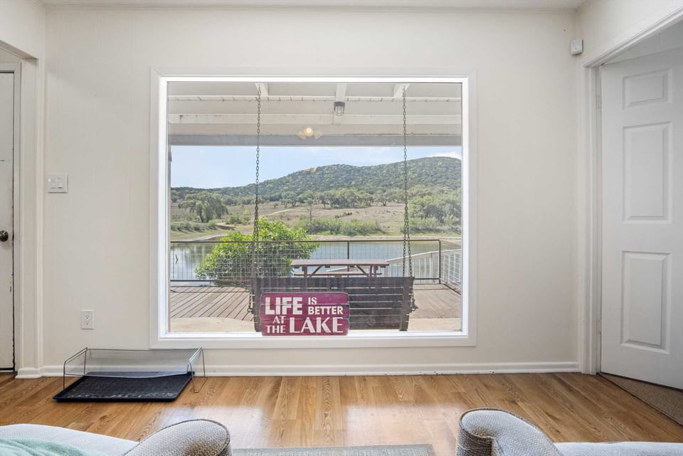 A large picture window perfectly frames the outdoor porch and lake view, accented by a charming "Life is Better at the Lake" sign that brings a touch of personality to the room.