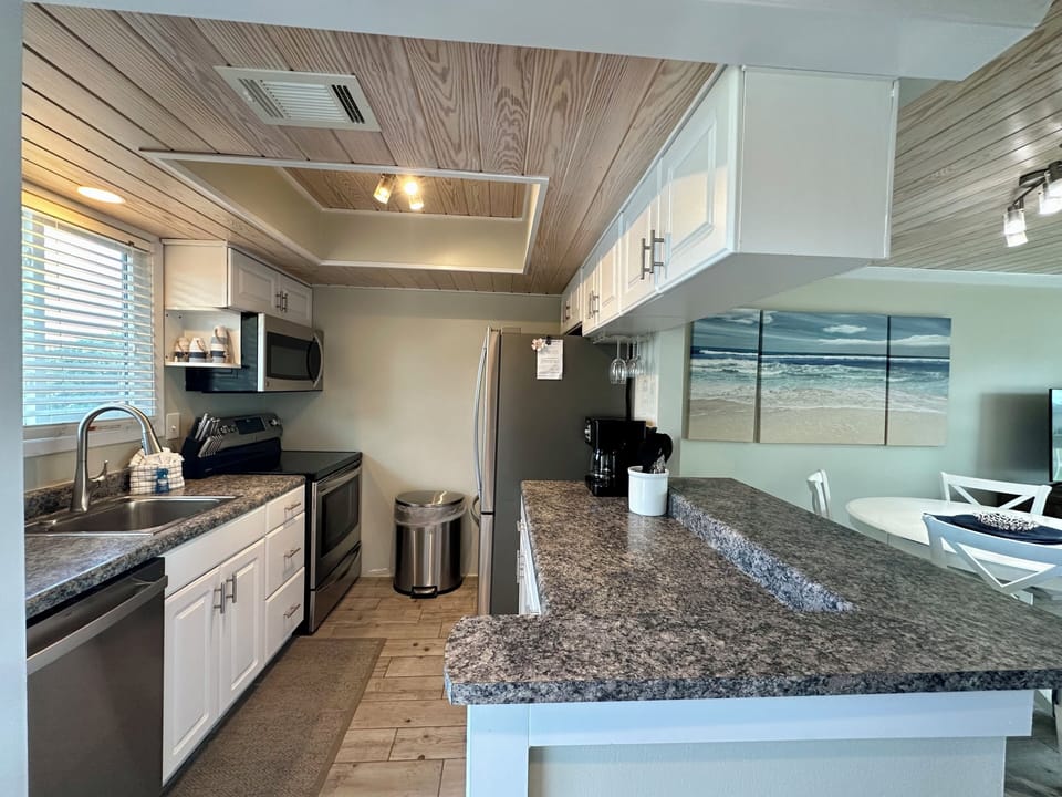 Your coastal kitchen blends modern elegance with warm wood ceilings, granite counters, and ocean artwork creating the perfect culinary retreat.