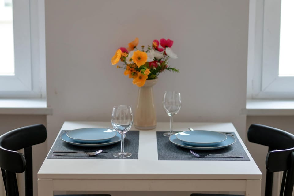 A dining table is set with plates and a vase of flowers as a decorative centerpiece. Soft tones create a welcoming dining atmosphere.