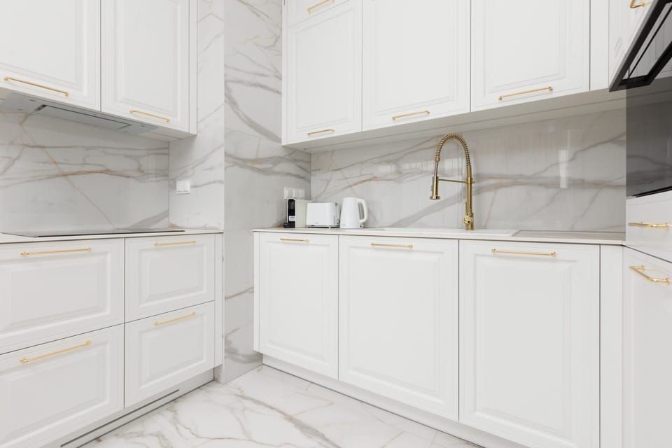 A sleek white kitchen with marble countertops, gold hardware, and built-in appliances.

