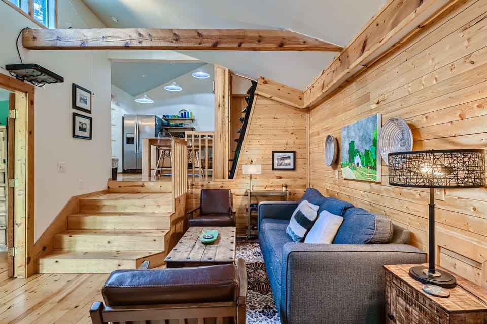A cozy wooden interior with an open loft design featuring a timber staircase, a gray sofa, a rustic coffee table, a lamp, and wall art. The kitchen area is visible in the background.