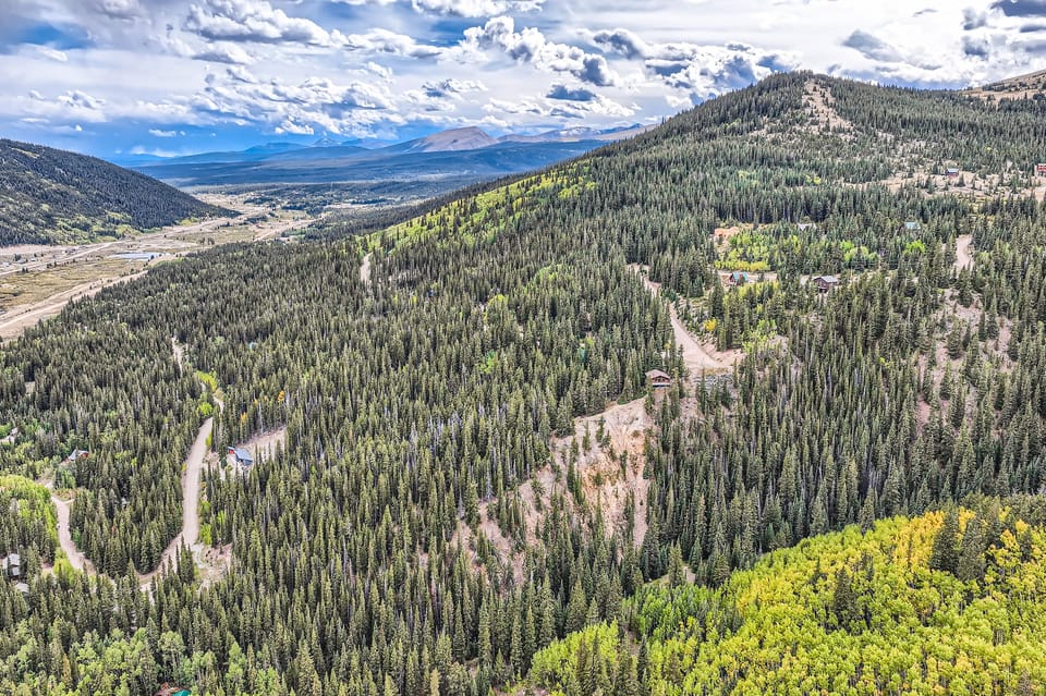 Aerial view of a mountainous landscape with winding roads, dense forests, and patches of open land under a partly cloudy sky.