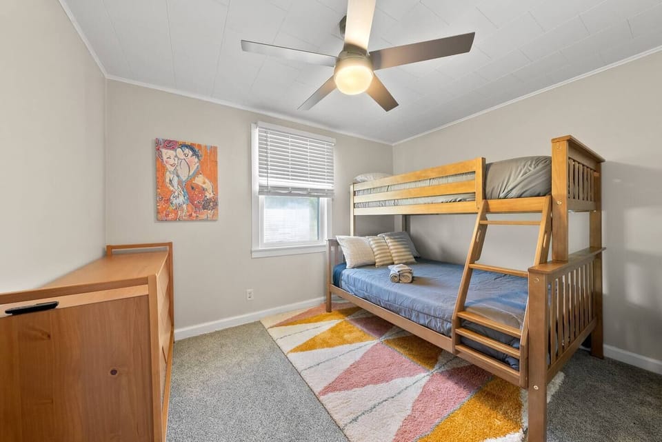 A colorful area rug adds a playful touch to the room’s decor. To the left, a wooden desk or dresser offers storage and functionality. The wall is adorned with an artwork featuring two characters, adding a personal and creative touch to the space. 