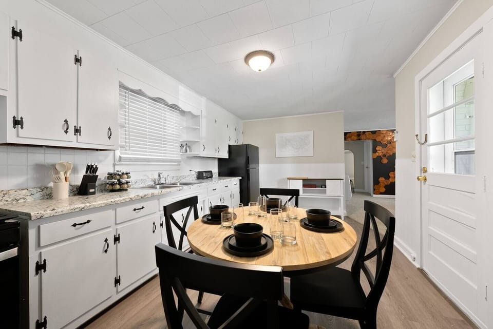 The kitchen is equipped with white cabinetry accented with black hardware, and a light gray countertop. The appliances are of stainless steel, adding a modern touch to the space. In the foreground, there’s a round wooden dining table.