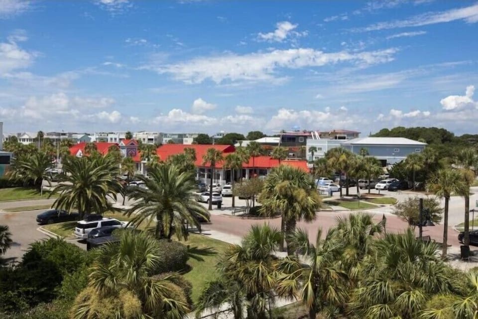 View of a parking lot with palm trees and buildings in the background under a blue sky with clouds.