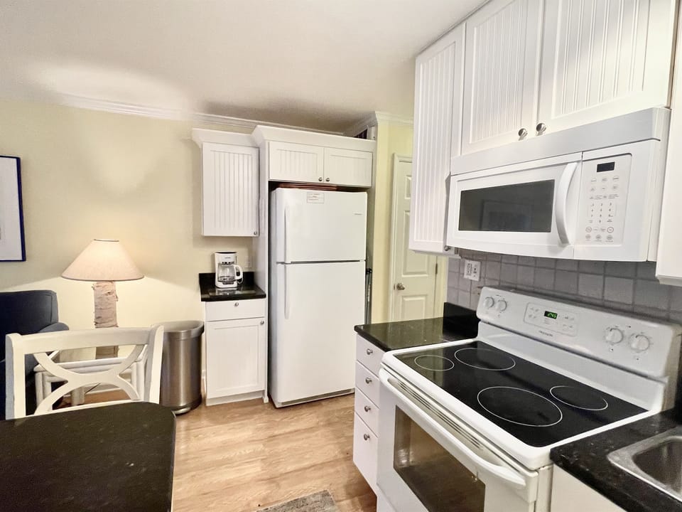 Compact kitchen with white cabinets, black countertops, refrigerator, stove, microwave, and coffee maker. A small dining area is visible with a wooden chair and lamp.