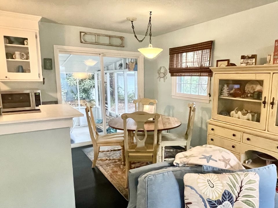 A cozy dining area with a round wooden table and four chairs near a window. A light blue couch with patterned pillows is in the foreground, and a hutch displays decorative items.