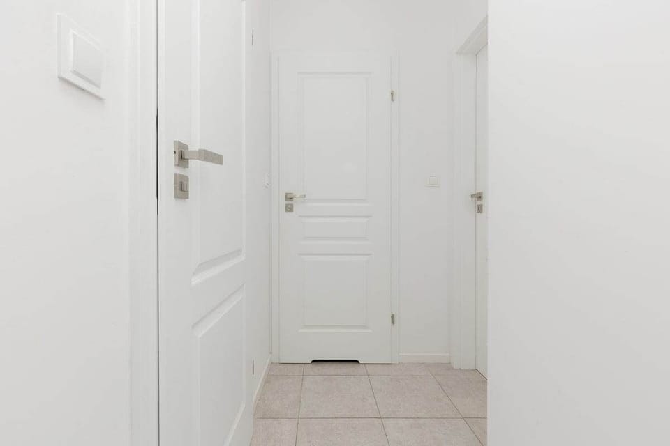 A hallway view, leading to a white door, with a tiled floor and bright walls, maintaining the apartment's minimalist style.
