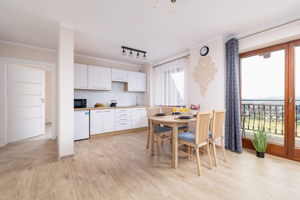 A spacious dining and kitchen area with wooden furniture, large windows, and access to a balcony with mountain views.
