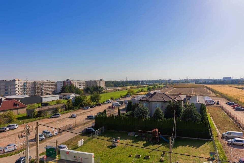 A panoramic outdoor view from the apartment, showcasing a green lawn, residential buildings, and open fields under a bright blue sky.






