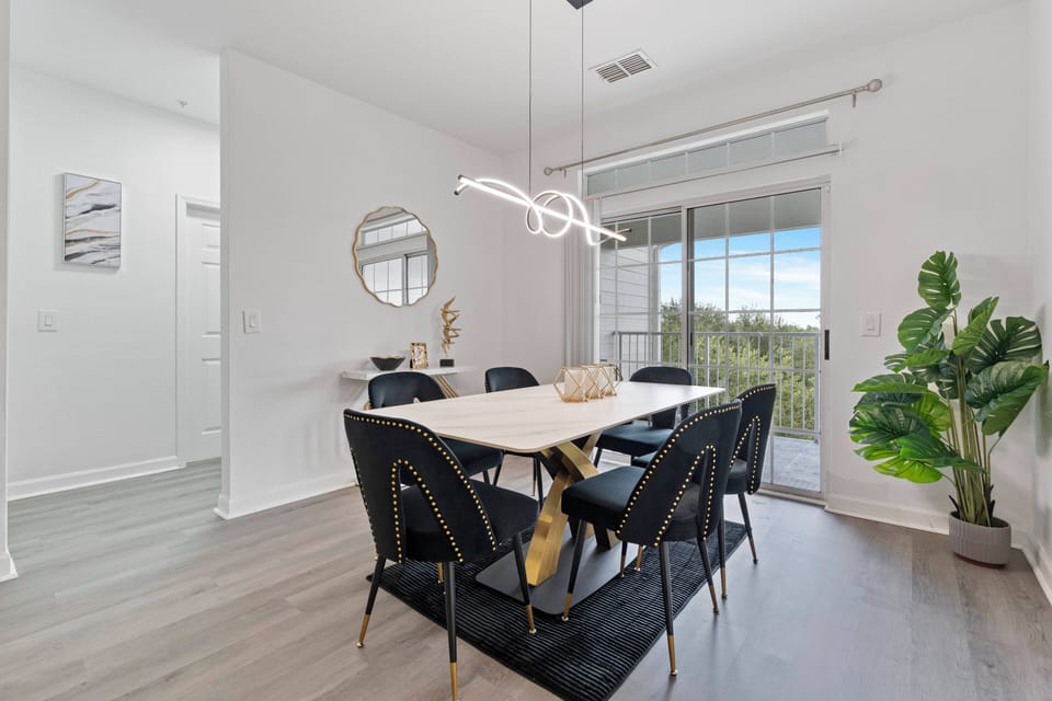 Enjoy the natural light flooding this chic dining area, where a stunning chandelier hangs above a sleek table with elegant black chairs. The soft area rug and fresh greenery create a relaxed, sophisticated ambiance for your meal.