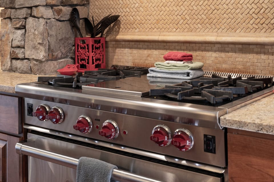 - Professional grade gas stove with red knobs and four burners for gourmet cooking - Handy utensils and folded dish towels keep everything within easy reach - Textured backsplash and stone accents complete the rustic kitchen aesthetic