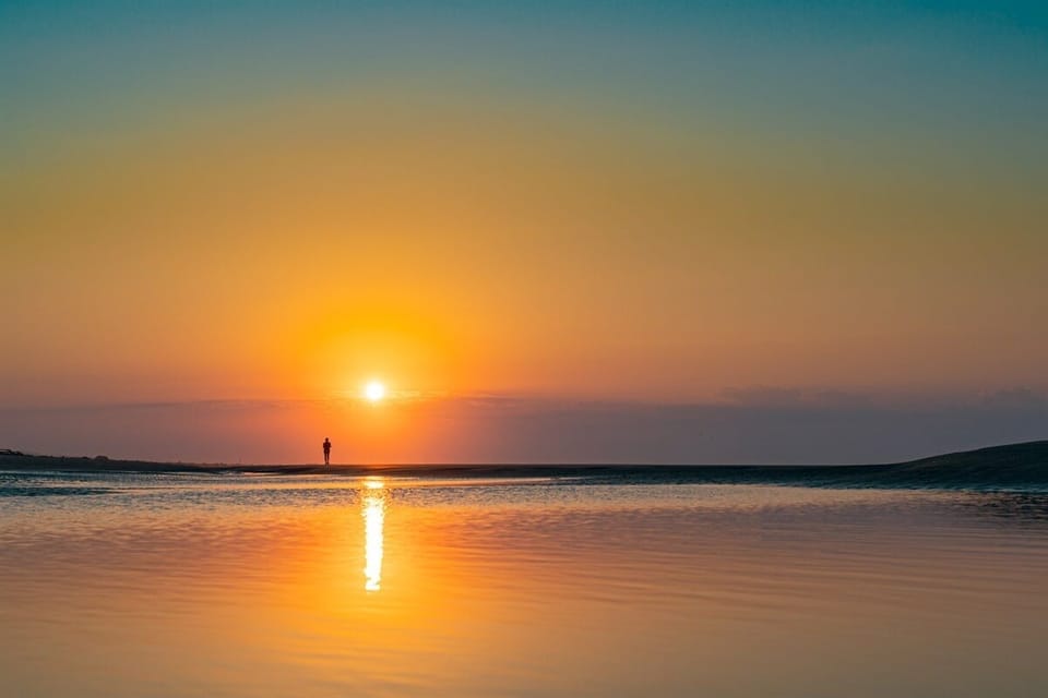 Person standing on a beach at sunset, with the sun low on the horizon, reflecting on the calm water.