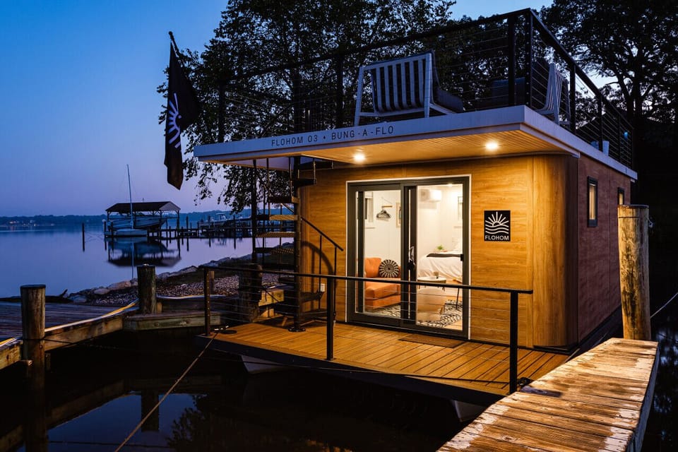 Bung-a-flo glowing at dusk at Ferry Point Marina on the Magothy River, with the FLOHOM flag flying and the cedar-clad exterior framing the cozy interior within.