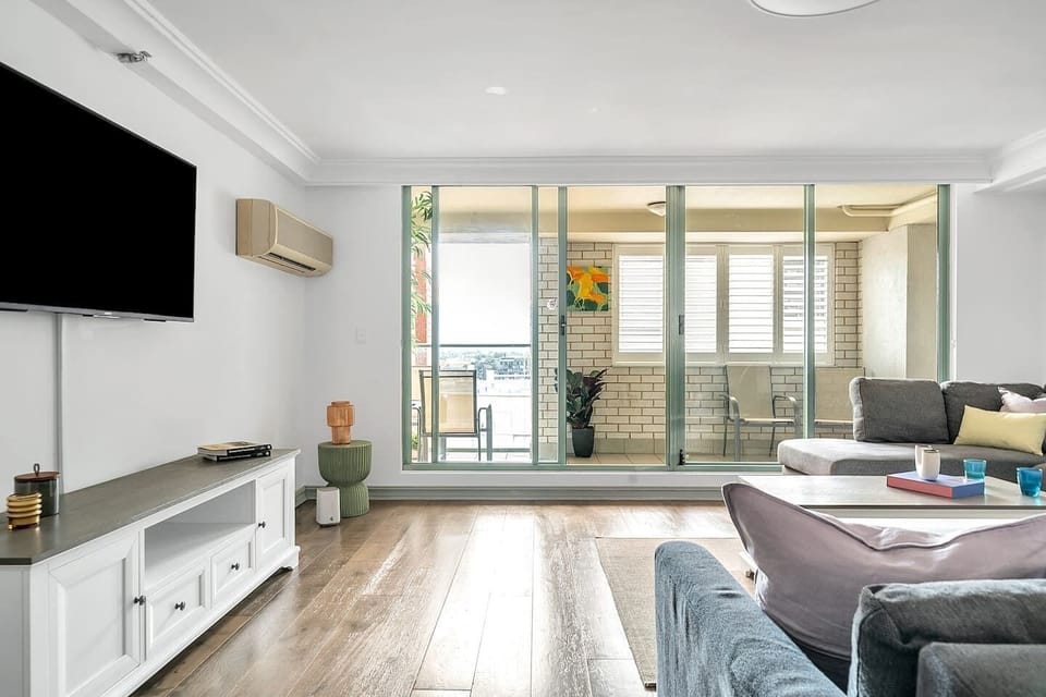 Unwind in the spacious living room, featuring a large HD TV mounted on the wall, extended lounge space, and ample storage. The living room seamlessly connects to the downstairs balcony, allowing you to enjoy the outdoors while relaxing indoors.